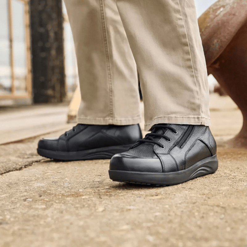 A man wearing beige trousers and black New Feet men's boots standing on a grey surface